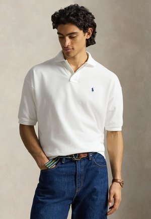 Jonge man met donker krullend haar die een wit poloshirt met marineblauw logo draagt, blauwe spijkerbroek, gestreepte riem en een bruin horloge, kijkt naar beneden.