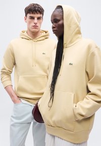 Deux mannequins portant des sweat-shirts à capuche beige avec poches avant et un logo crocodile vert, debout devant un fond uni clair.