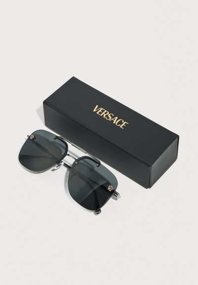 Γυαλιά ηλίου με μαύρους τετράγωνους φακούς και ασημένια πλαίσια, τοποθετημένα δίπλα σε μαύρη θήκη Versace πάνω σε απλή λευκή επιφάνεια.