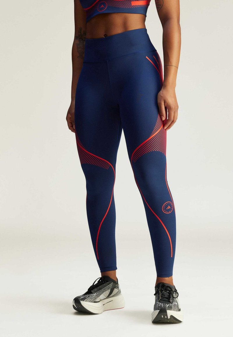 Sotilasnavy- ja punaiset urheiluleggingsit, jotka on valmistettu joustavasta kankaasta, jossa on kaarevat saumat. Leggingsit sisältävät logoyksityiskohtia ja kontrastivärisiä viivoja sivuilla.