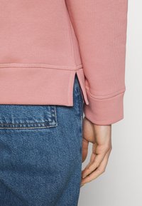 Sudadera de color rosa claro con cuello redondo, puños acanalados y aberturas laterales. Usada con jeans de mezclilla azul, que muestran una textura de tela y un diseño casual.