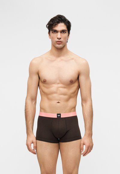 Muskulöser Mann mit dunklen, lockigen Haaren, der dunkelbraune Boxershorts mit einem pinken Bund und Markenlogo in der Mitte trägt, vor einem grauen Hintergrund steht.