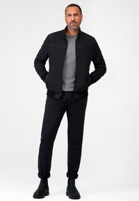 Schwarze Bomberjacke mit Stehkragen, Reißverschluss und strukturierten Details, kombiniert mit einem grauen Hemd und schwarzen Jeans mit umgeschlagenem Saum. Schwarze Chelsea Boots.