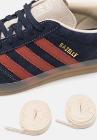 Marinblå mockasko med röda läderremmar, guld "Gazelle"-text och en klar gummisula. Inkluderar två krämfärgade skosnören.