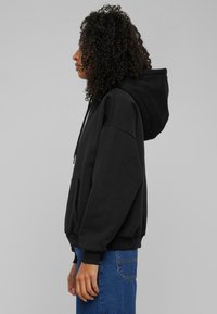 Svart hettegenser med frontlomme, snor og kenguru-stil. Har en avslappet passform og ribbestrikkede cuffs, kombinert med blå jeans.