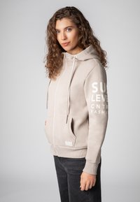 Licht beige zip-up hoodie gemaakt van zachte stof, met een capuchon met trekkoord, kangoeroezakken en witte tekst op de mouw.