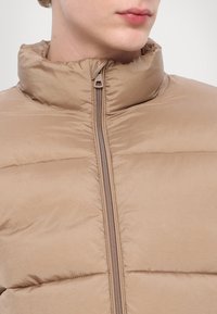 Jachetă bej căptușită cu guler înalt și textură netedă. Are un fermoar frontal și un model cusut orizontal.