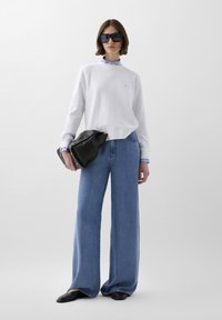 Hvid sweatshirt med ribbet halsudskæring, lyseblå stribede skjorteærmer, brede denimjeans, sort oversized håndtaske og sorte flade sko.