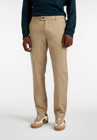 Pantaloni chino beige con taglio dritto, dotati di chiusura con bottone e tasche frontali, indossati con sneakers di colore chiaro.