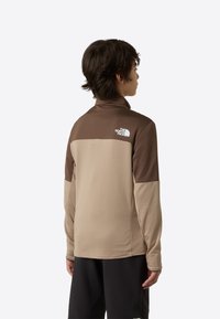 Tricou sport maro și bej cu mâneci lungi, guler înalt, textură nervurată și logo-ul The North Face pe partea superioară a spatelui.