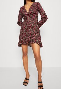 Schwarzes Blumenkleid mit langen Ärmeln, V-Ausschnitt, tailliertem Schnitt und geschichteten Rüschensaum. Es hat kleine, mehrfarbige Blumen auf dem gesamten Kleid.