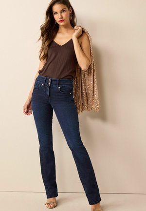 Jeans svasati in denim scuro con vita alta, due tasche frontali e chiusura con bottone, abbinati a un top a maniche corte marrone a costine e un poncho luccicante color oro rosa.