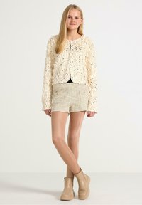 Hæklede cremefarvede cardigan med intrikat tekstur, sorte knapdetaljer, parret med beige tweed-shorts og beige ankelstøvler.