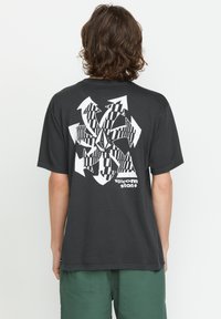 Donkergrijze t-shirt met korte mouwen, voorzien van een groot wit abstract grafisch ontwerp en de tekst "Volcom Stone" onderaan. Soepele katoenen textuur.