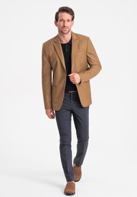 Ruskea bleiseri mustan poolopaidan päällä, kapeat harmaat housut ja ruskeat loaferit. Suuressa tekstuurissa on yksi tasku yksityiskohtana bleiserissä.