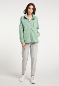Giacca in pile verde chiaro con colletto alto, zip sul davanti e tasca sul petto; abbinata a jeans grigi chiari e sneakers bianche.