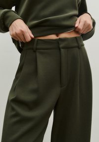 Pantalons olive vert taille haute en tissu doux, avec un design plissé et des passants de ceinture, dotés de poches latérales visibles.