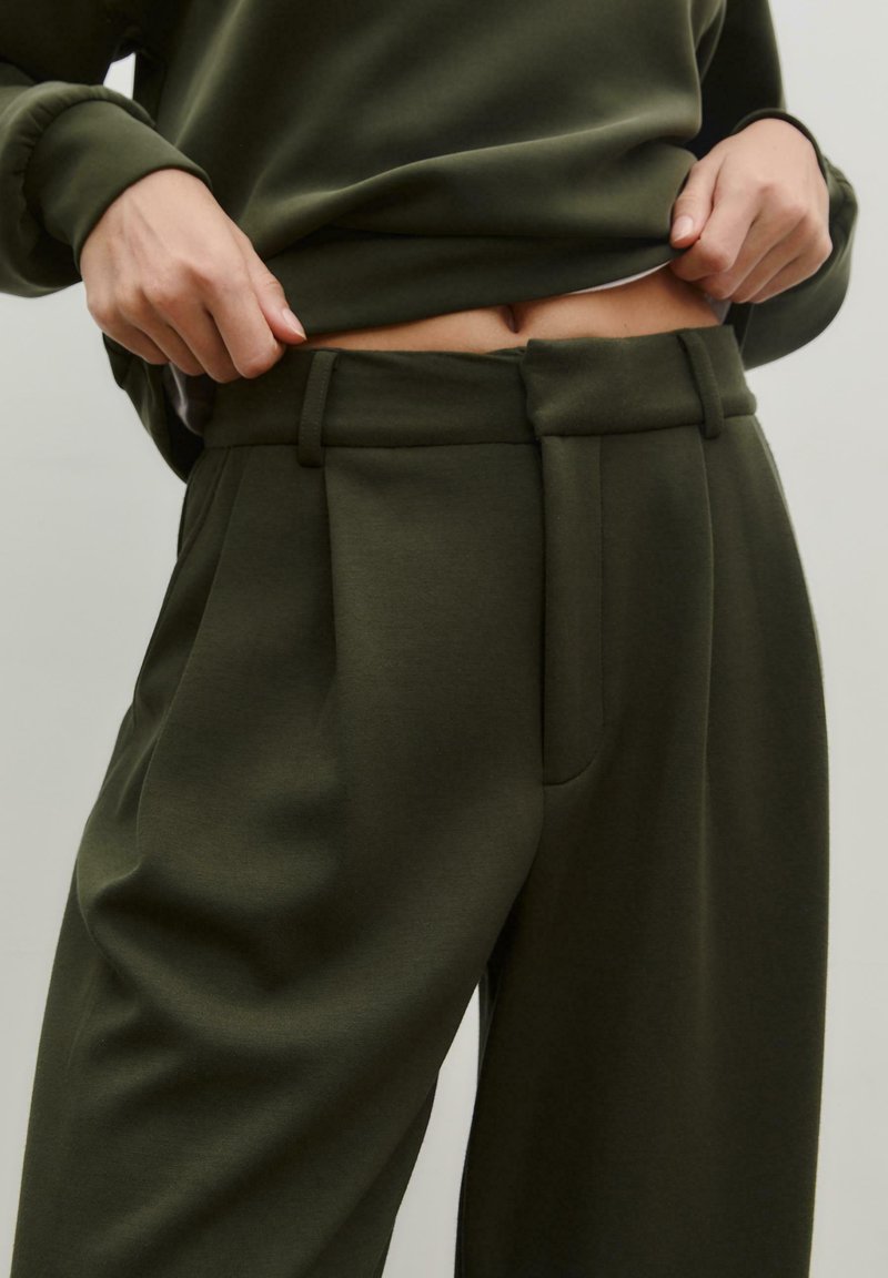 Pantalons olive vert taille haute en tissu doux, avec un design plissé et des passants de ceinture, dotés de poches latérales visibles.