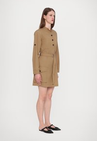 Abito button-up color tan con maniche lunghe, due tasche frontali, vita aderente con cintura, e scarpe nere slip-on con punta affusolata.