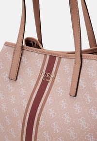 Vaaleanpunainen synteettinen kassi, jossa on monogrammikuosi, kultainen logotehoste ja punainen pystysuora raita. Suuri sileä pinta ja nahkamaiset hihnat.