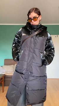 Osoba ubrana w długi czarny puchowy płaszcz, okulary przeciwsłoneczne z pomarańczowymi szkłami oraz torbę na ramię, stoi w pomieszczeniu z rękami w kieszeniach płaszcza.