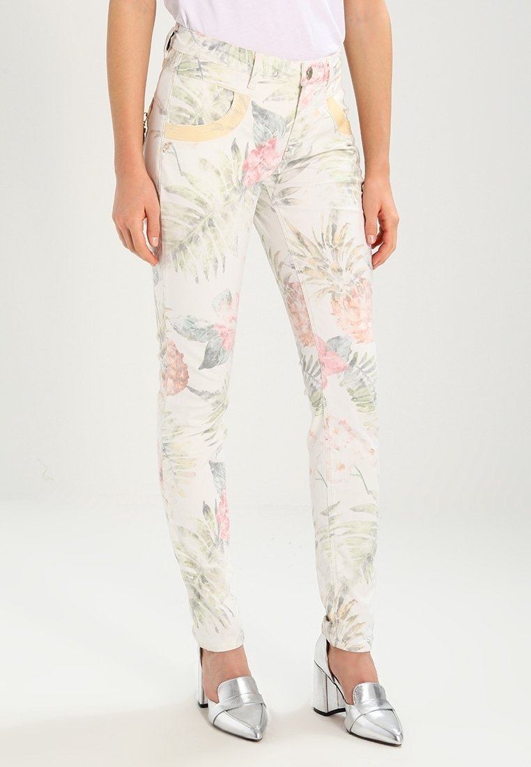 Femme portant un pantalon blanc ajusté avec un imprimé floral tropical pastel et des détails de poche jaunes, associé à des chaussures à talons blocs argentées.
