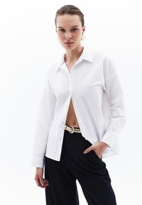 Witte blouse met lange mouwen, gemaakt van een gladde stof, met een puntige kraag en een open voorkant, gecombineerd met zwarte broek en een gouden riem.