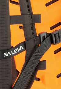 Arnés de tela naranja con correas negras, hebilla ajustable y logo. Superficie texturizada con recortes de ventilación para una mejor transpirabilidad.
