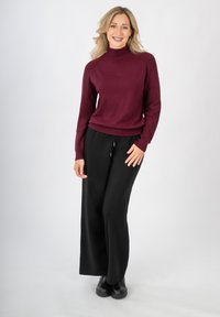 Weinrotem Rollkragensweater mit gerippten Bündchen, kombiniert mit weit geschnittenen schwarzen Hosen. Das Model trägt schwarze Knöchelstiefel und präsentiert ein entspanntes Outfit.
