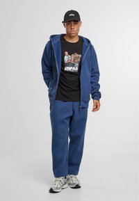 Blaues Zip-Hoodie über einem schwarzen T-Shirt mit Grafikdruck, kombiniert mit passenden blauen Jogginghosen und grauen Sneakers.