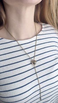 Aranyszínű nyaklánc kör alakú medállal, letisztult kialakítással, texturált láncon lóg, csíkos ing fölött viselve.