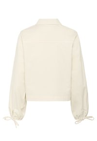 Crèmekleurige korte jas met lange, opgezette mouwen. Kenmerkend zijn de puntige kraag en striksluitingen bij de manchetten. Vloeiend materiaal met minimale details.