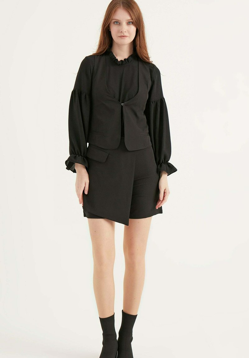 Zwarte mouwloze jack met V-hals en voorzakken, gecombineerd met zwarte, op maat gemaakte shorts. Model draagt zwarte ribbeltekkousen.