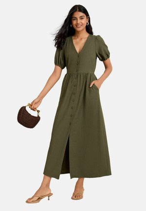 Abito verde oliva, fino al ginocchio, con maniche a sbuffo, scollatura a V, abbottonatura frontale e tasche laterali. Indossato con sandali beige e una borsa intrecciata.