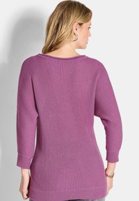 Frau mit blonden Haaren, die Creolen und einen lila gerippten Pullover trägt, von hinten vor einem weißen Hintergrund gezeigt.