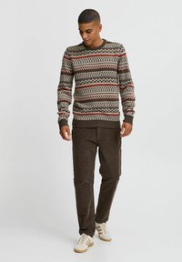 Pulover tricotat maro cu modele și desene roșii și crem, asociat cu pantaloni din catifea corduroy maro închis și adidași albi.
