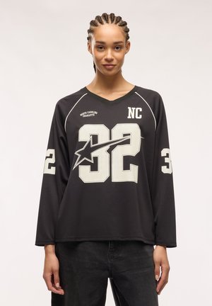 Giovane donna con capelli intrecciati che indossa una maglia sportiva nera a manica lunga con il numero 32 e il testo "NC" e "North Carolina Charlotte".