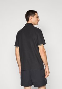 Sort polo-shirt med krave, korte ærmer og en lidt løs pasform, parret med mørke shorts. Viser synlige tatoveringer på armen.
