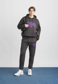 Juodas gobtuvas ir sportinės kelnės su violetiniu „Reebok“ logotipu. Minkšta medžiaga, laisvas kirpimas. Baltos sportbačiai su įvairiaspalviais akcentais. Paprasta fona.