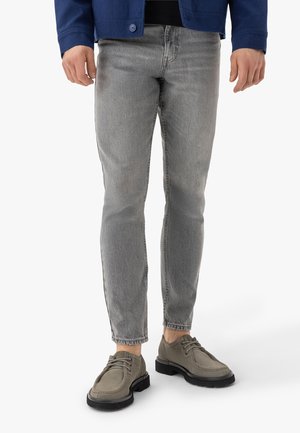 Mężczyzna ubrany w jasnoszare obcisłe jeansy, szare sznurowane buty i ciemnoniebieską kurtkę, stojący na białym tle.