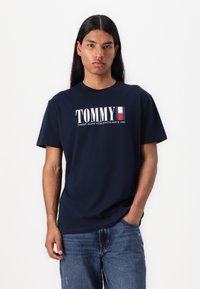 Marineblauwe katoenen T-shirt met opvallende witte "TOMMY"-tekst en een rood, wit en blauw rechthoekig logo. Relaxte pasvorm, ronde halsontwerp.