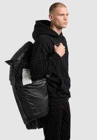 Chaqueta acolchada negra con textura brillante y acolchonada, usada sobre una sudadera con capucha negra. Cuenta con cierre de cremallera y una etiqueta visible en el interior de la chaqueta.