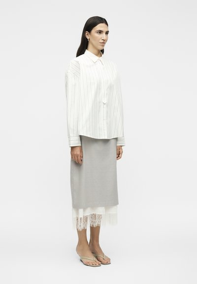 Γυναίκα με μακριά σκουρόχρωμα μαλλιά φοράει ένα λευκό πουκάμισο με ρίγες, γκρι μίντι φούστα με λευκή δαντέλα στο τελείωμα και μπεζ πέδιλα με ανοιχτά δάχτυλα, στέκεται πλάγια.