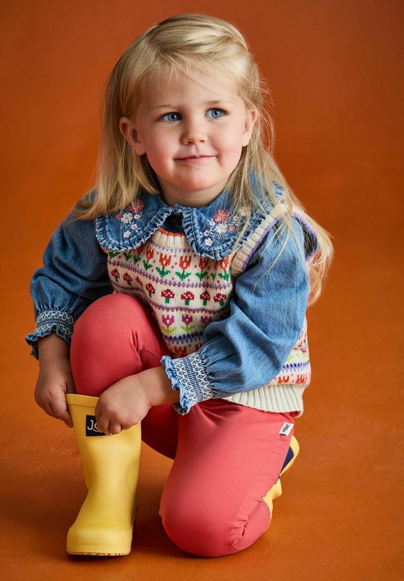 Ett ungt barn med blont hår som bär en blommig denimkrage skjorta, mönstrad stickad väst, korallfärgade byxor och gula gummistövlar på knäna på en orange golv.