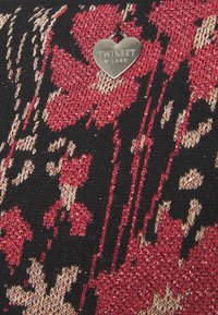 Pendentif en argent en forme de cœur gravé de « TWINSET MILANO » sur un tissu tricoté aux motifs rouges, noirs et beiges.