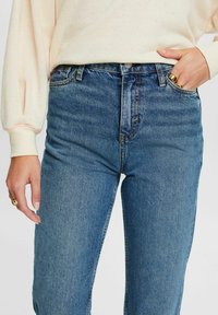 Jeans de mezclilla azul de cintura alta con corte recto, costuras visibles y detalle de bolsillo; combinados con un sudadera clara y texturizada en color crema.