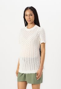 Maglione a maniche corte lavorato a maglia di colore bianco con motivo a rombi traforati, colletto rotondo e orlo a coste. Abbinato a pantaloni corti verdi.