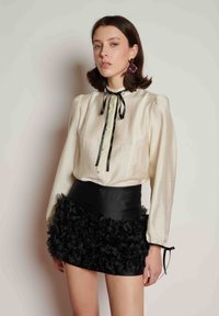 Κρεμ μπλούζα με υφή, φουσκωτά μανίκια, μαύρη κορδέλα στη λαιμόκοψη, κούμπωμα με κουμπιά, και μαύρη σατέν μίνι φούστα με βολάν στο τελείωμα.