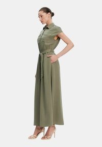Abito maxi verde oliva con maniche corte, vita cintata e tasche laterali. Il tessuto appare liscio con una vestibilità ampia e un colletto strutturato.