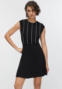 Abito nero in maglia con corpetto aderente, maniche corte e gonna svasata. File verticali di perle adornano il corpetto, aggiungendo dettagli.